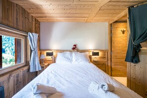A wooden-paneled room with a bed, a window with curtains, a nightstand with a lamp, and a door with a coat hanging on it. - Cocon des Aravis - Beau duplex avec balcon (Le Grand Bornand)