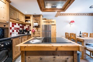 A kitchen with wooden cabinets, a black countertop, a stainless steel refrigerator, and a dining area with wooden chairs and a table. - Cocon des Aravis - Beau duplex avec balcon (Le Grand Bornand)