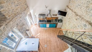 A high-ceilinged room with a stone wall, a wooden floor, a kitchen area with teal cabinets, and a dining table with chairs. - Les Magnans - Grospierres (Grospierres)