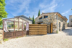 A stone building with a wooden gate, a glass structure with curtains, and a gravel driveway. - Le Cocon - Grospierres (Grospierres)
