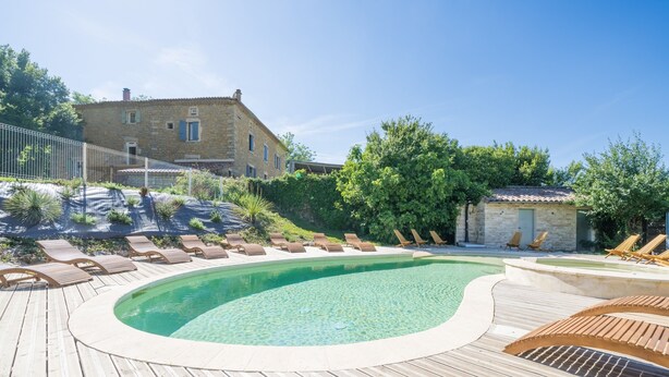 A wooden deck with a swimming pool, lounge chairs, and a stone building in the background. - Le Cocon - Grospierres (Grospierres)