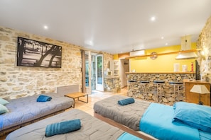 Une pièce avec deux lits, un mur en pierre, un coin cuisine et une vue sur l’extérieur par une porte ouverte.