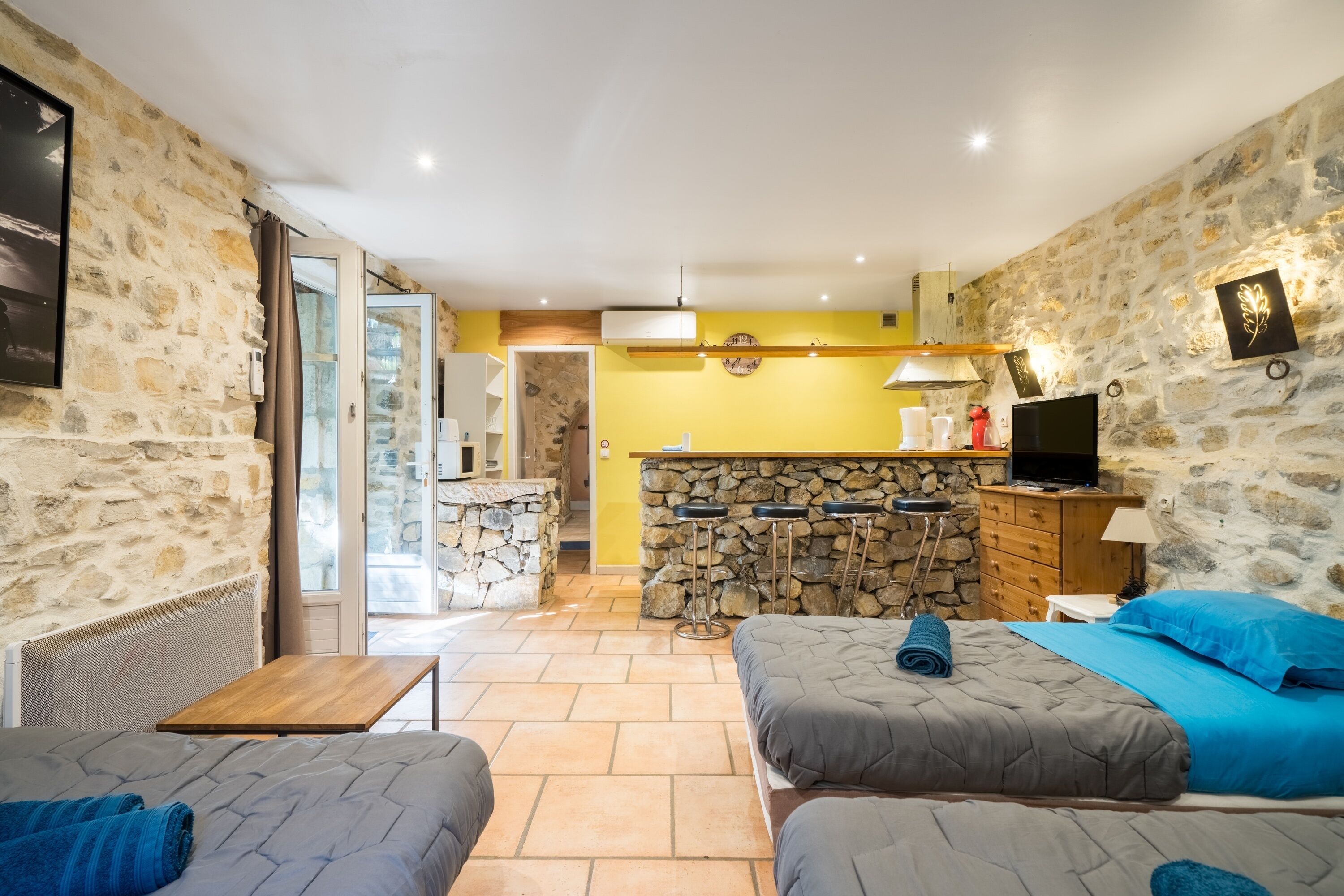 A room with two beds, a stone wall, a kitchen area, and a view through a glass door.