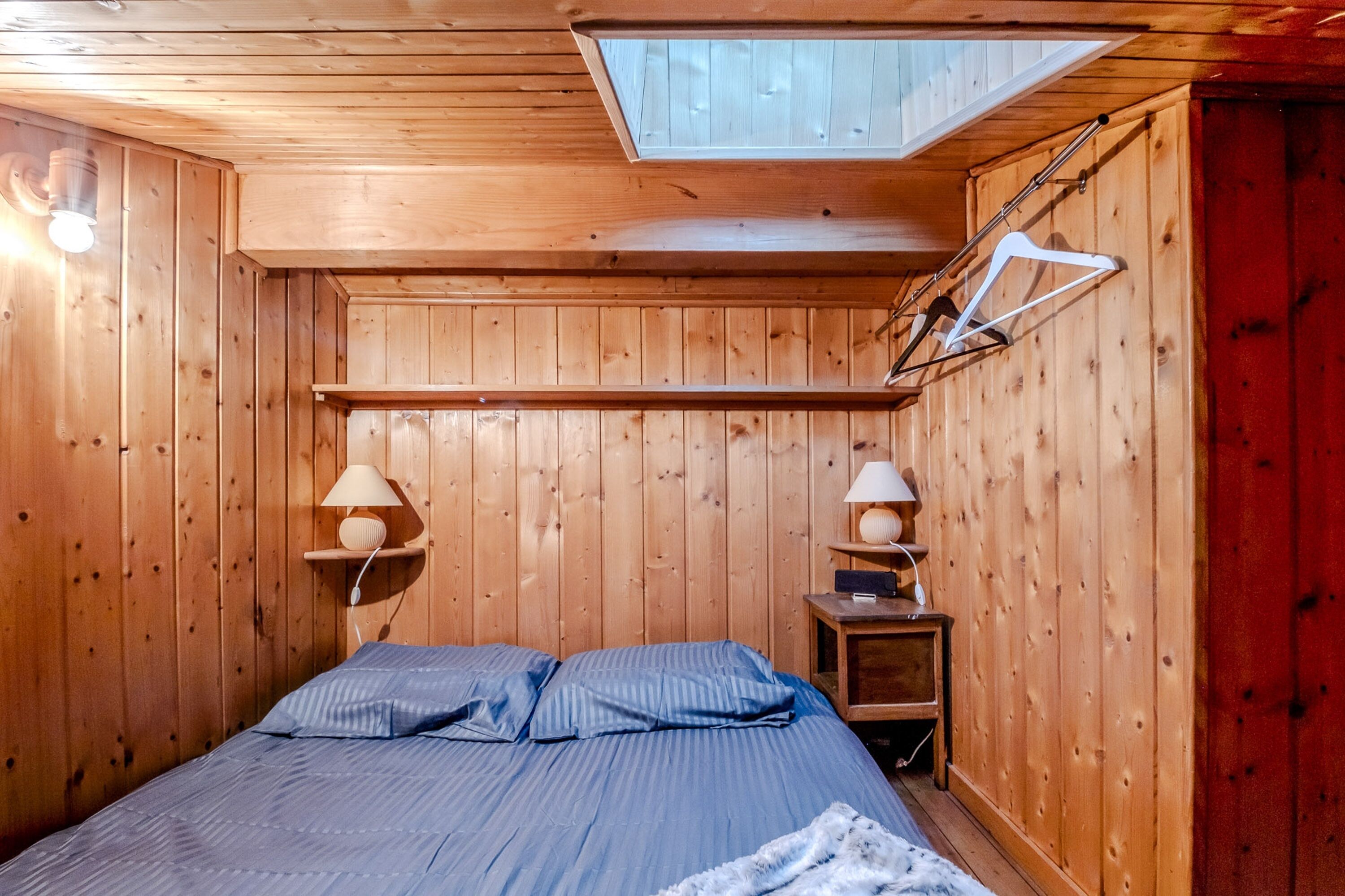 A wooden room with a bed, two bedside lamps, a skylight, and a red wall.