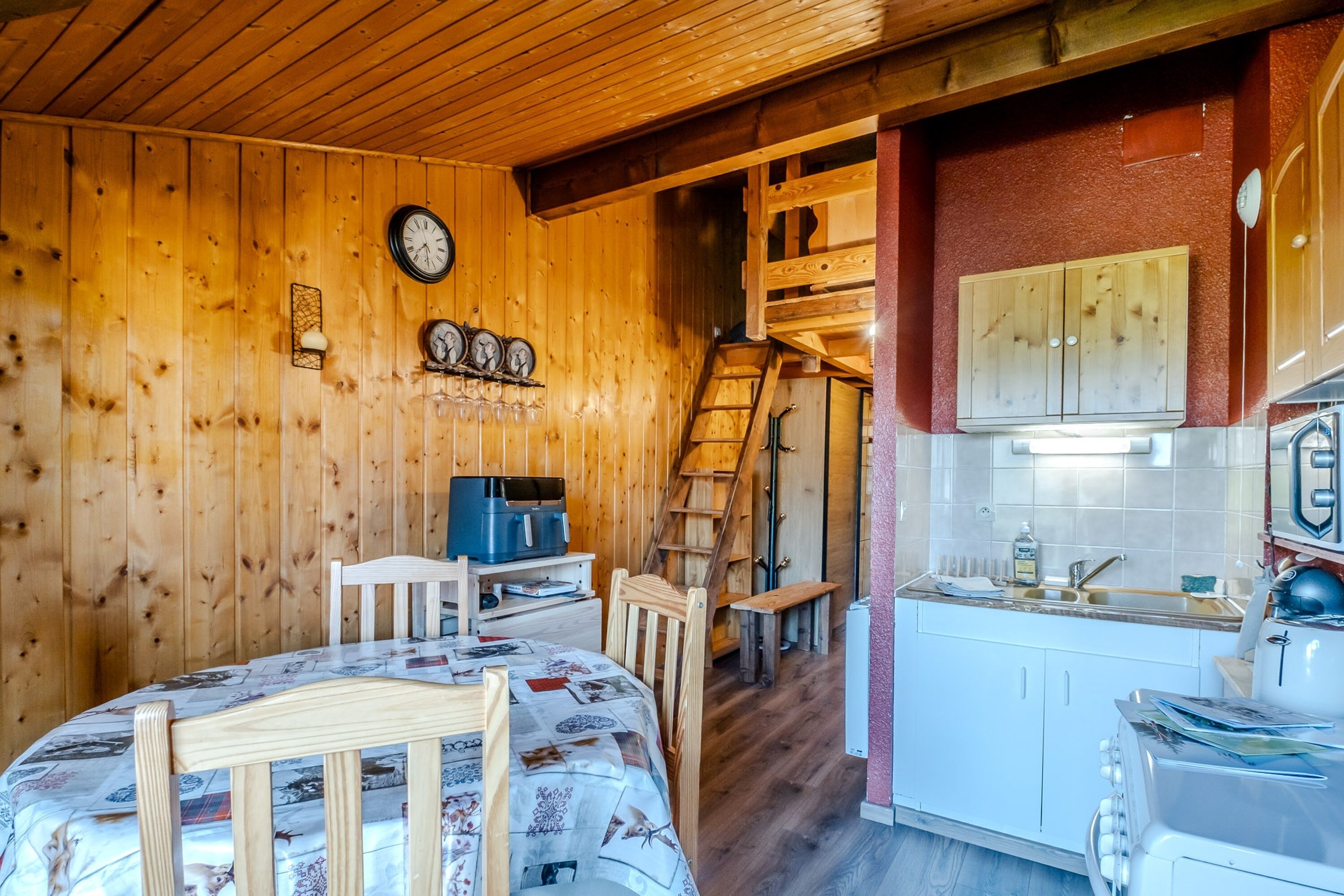 Interior de una cabaña de madera con una mesa de comedor, sillas, una cocina con armarios blancos, una escalera de madera y un reloj en la pared.