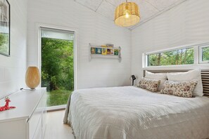 A bedroom with a large bed, a bedside table, a lamp, a shelf with books, and a window with a view of greenery. - Holiday home with large doors and forest (Gilleleje)