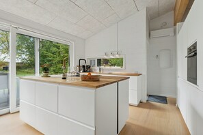 A modern kitchen with a large island, white cabinetry, and a view through the glass door. - Holiday home with large doors and forest (Gilleleje)