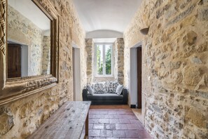 A stone-walled corridor with a wooden bench, a sofa with patterned cushions, and a large mirror on the left. - Le Mas - Grospierres (Grospierres)