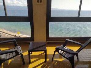 Ett rum med stora fönster med utsikt över en strand, två plaststolar och ett litet bord.