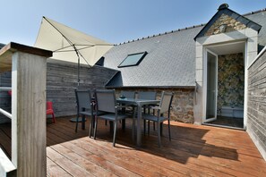 Een houten terras met een tafel, stoelen, een klein raam en een deur naar een kamer met een plant.