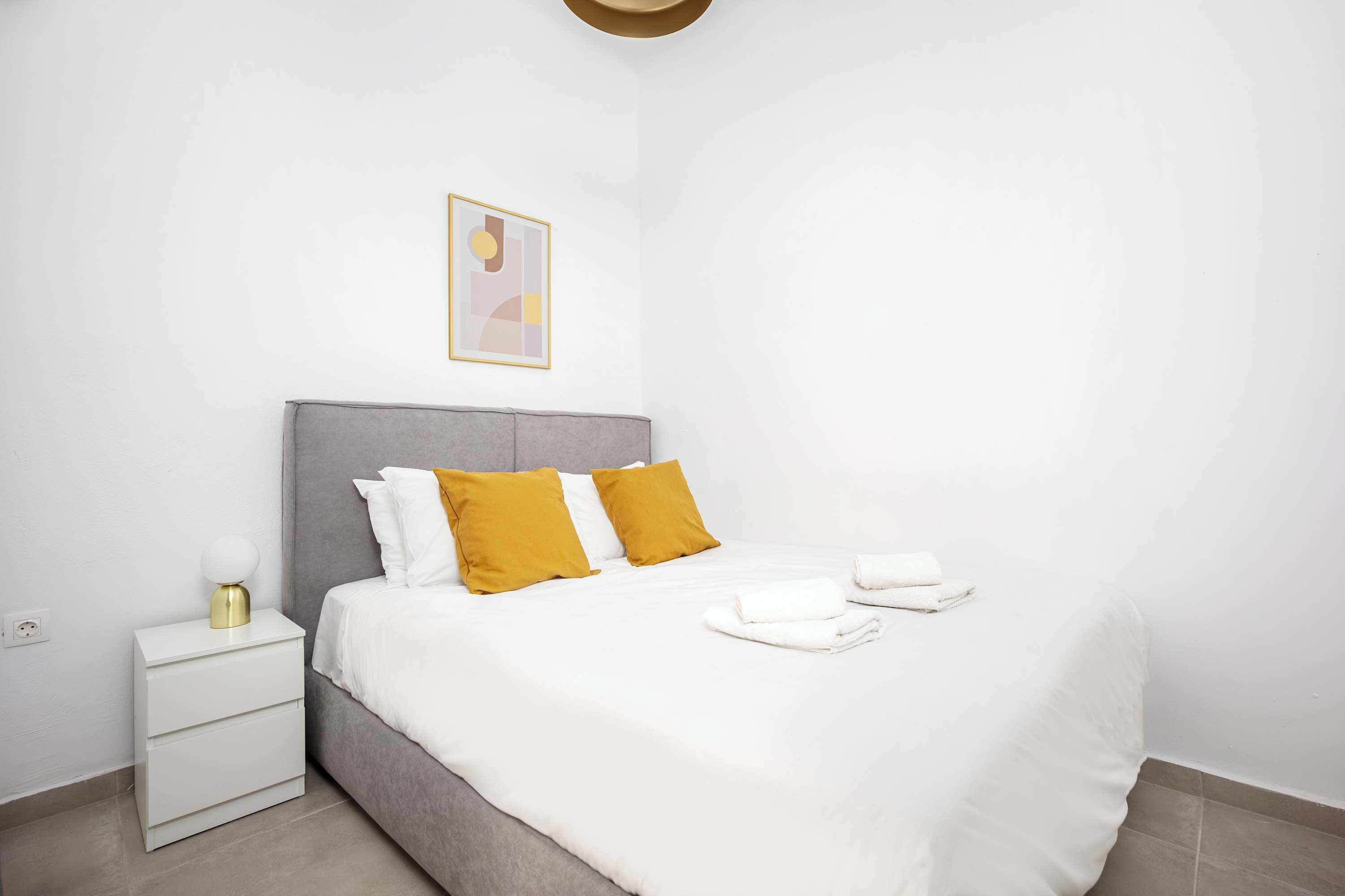 A neatly made bed with white bedding and two mustard-colored pillows, a bedside table with a lamp, and a wall-mounted abstract art piece.