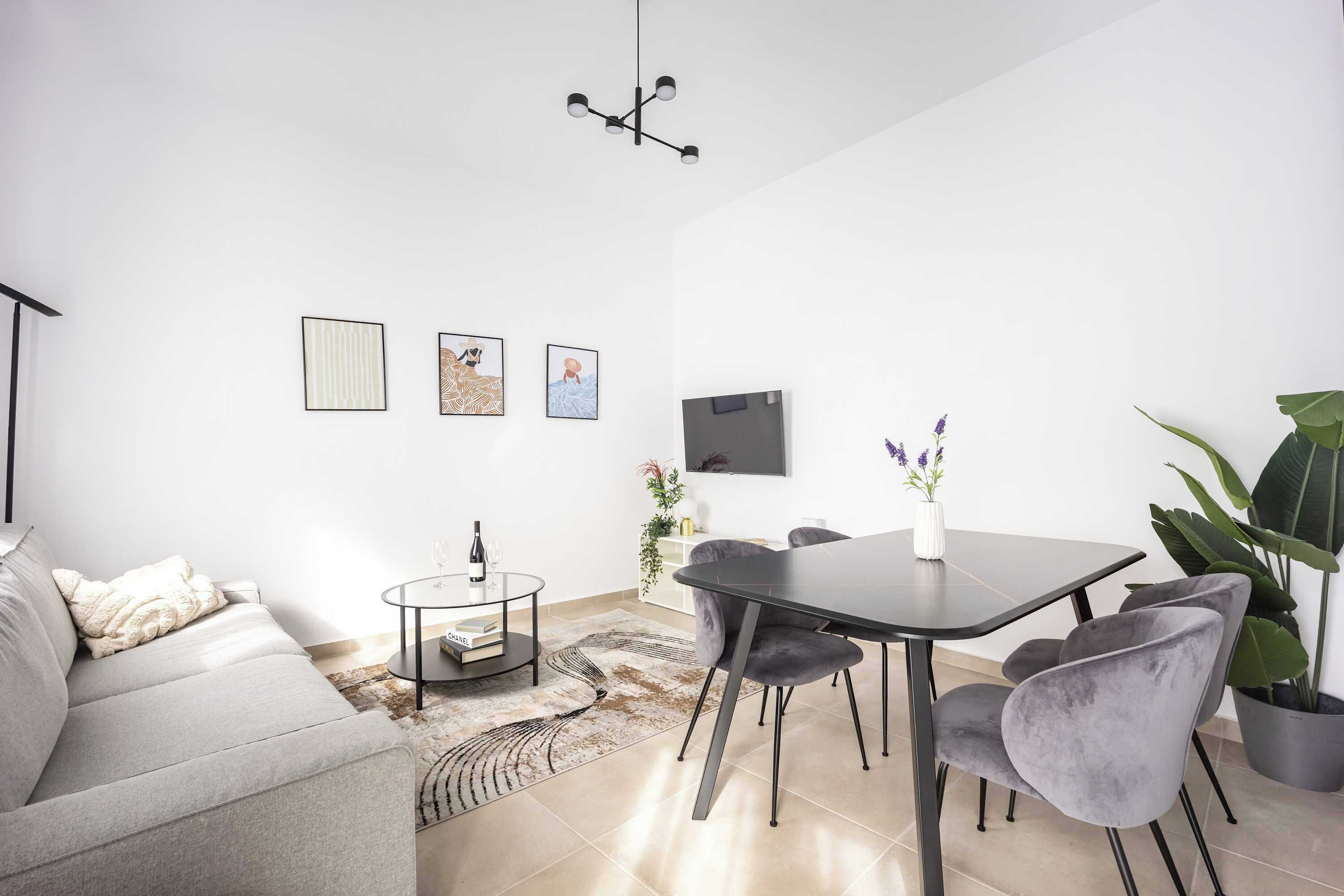 A modern living room with a sofa, a round coffee table, a dining table with chairs, a flat-screen TV mounted on the wall, and a large potted plant.