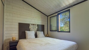 Quarto com teto de madeira, uma cama grande com cabeceira em macramé, duas mesas de cabeceira com abajures e uma janela com vista para árvores.