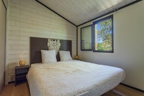 Ein Schlafzimmer mit Holzbalkendecke, einem großen Bett mit Makramee-Kopfstück, zwei Nachttischen mit Lampen und einem Fenster mit Blick auf Bäume.