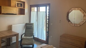 A room with a bed, a chair, a table, a dresser, an air conditioning unit, a window with curtains, and a wall-mounted mirror.