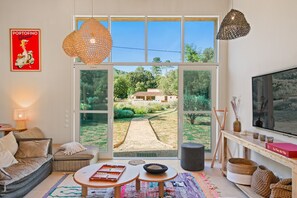 A modern living room with a large sliding glass door leading to a garden, a sofa, a coffee table, and a TV. - Les 3 thyms - Avec Piscine Privée (Méounes-lès-Montrieux)