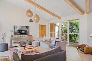 En moderne stue med sofa, sofabord og fjernsyn. Der er træbjælker i loftet, et stort vindue og et træskab.