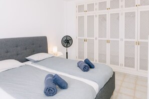 A bedroom with a grey upholstered headboard, a bed with grey linens, two rolled towels, a bedside table with a lamp, and a white wardrobe with perforated panels. - Bright 1-Bedroom Apartment with Private Terrace and Views | Pool (Playa de las Américas)