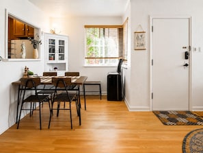 Un espacio habitable pequeño con zona de comedor, una cocina compacta, un ventanal con persianas, una puerta con cerradura y un pequeño purificador de aire.