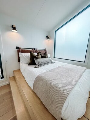 Ein modernes Schlafzimmer mit einem großen Bett, einem Holz-Kopfteil, zwei Wandleuchten und einem großen Fenster.