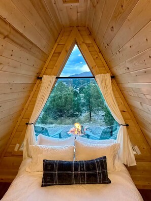 Eine Holzhütte mit einem Dreiecksfenster, einem Bett mit Kissen und Blick auf Wald und Berge.