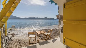 An outdoor seating area with two wooden chairs and a table, overlooking a beach with a clear sky and distant mountains.