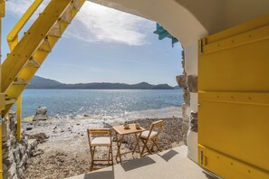 An outdoor seating area with two wooden chairs and a table, overlooking a beach with a clear sky and distant mountains. - Ky-ma Boat House (Milos)