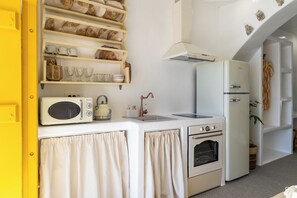 A compact kitchen with a microwave, oven, and refrigerator, a sink, and a shelf with dishes and glasses.