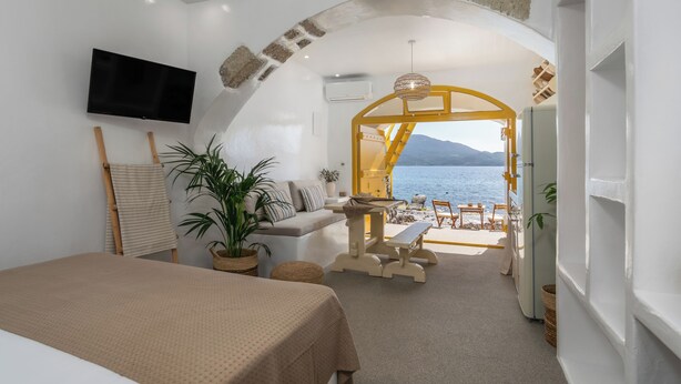 A room with a bed, a TV, a sofa, a table, and a bench. There is a view of the sea and mountains through an open archway.