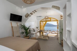 A room with a bed, a TV, a sofa, a table, and a bench. There is a view of the sea and mountains through an open archway. - Ky-ma Boat House (Milos)