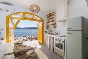 A room with a bed, a small table with a bowl of fruit, a kitchen area with a refrigerator, oven, and microwave, and a view of the sea through an open archway.