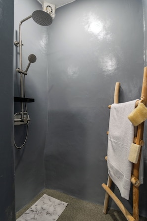 A small, narrow bathroom with a shower, a wooden drying rack with towels, and a patterned mat on the floor.