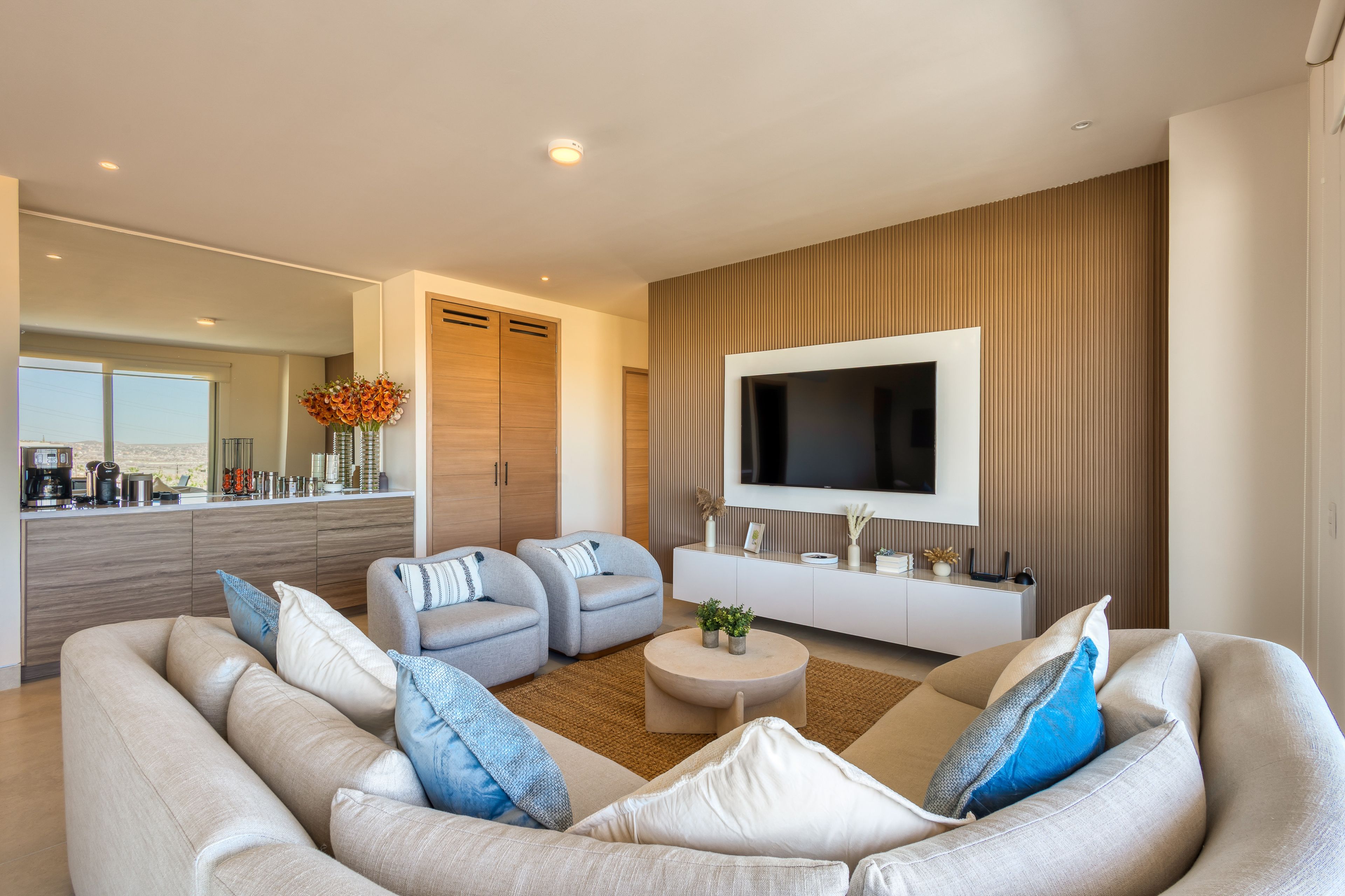 A modern living room with a large sofa, two armchairs, a coffee table, and a flat-screen TV mounted on the wall.