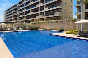 A swimming pool with lounge chairs and a building with balconies in the background. - Bright Coastal Condo With Ocean-view Balcony, Gym, Pools, BBQ & Kids Park (Cabo San Lucas)