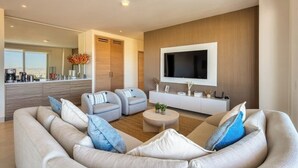 A modern living room with a sofa, two armchairs, a coffee table, a TV, and a kitchen area with a view. - Bright Coastal Condo With Ocean-view Balcony, Gym, Pools, BBQ & Kids Park (Cabo San Lucas)
