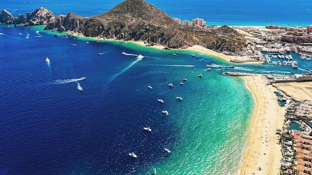 An aerial view of a coastal area with a prominent hill, clear blue waters, and a marina with boats. - Bright Coastal Condo With Ocean-view Balcony, Gym, Pools, BBQ & Kids Park (Cabo San Lucas)