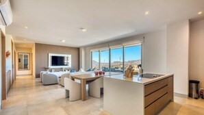 A modern kitchen with a large island, a dining area with a table and chairs, a TV mounted on the wall, and a view of the cityscape through large windows. - Bright Coastal Condo With Ocean-view Balcony, Gym, Pools, BBQ & Kids Park (Cabo San Lucas)