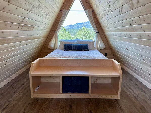 Une chambre mansardée en bois, avec un lit, une vue sur la verdure et les montagnes, et un plafond en pente.