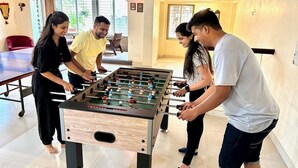 Un groupe de personnes joue au baby-foot dans une pièce dotée d’une grande fenêtre, d’une table à manger et d’une chaise rouge.