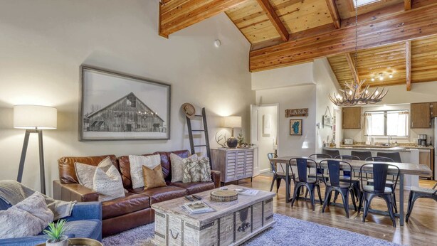 Een ruime woonkamer met een bruine leren bank, een salontafel, en een eethoek met stoelen, met een keuken op de achtergrond.