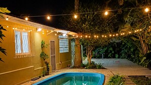 A swimming pool with a tiled deck, surrounded by greenery and a building with string lights.