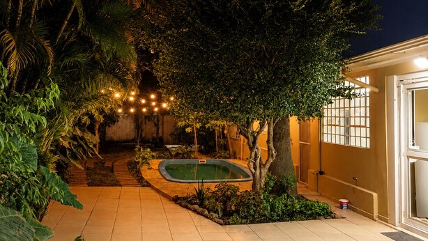 A well-lit outdoor area with a swimming pool, surrounded by lush greenery and a tiled patio.