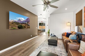 Ein modernes Wohnzimmer mit großem, an der Wand montierten Fernseher, Deckenventilator, Sofagarnitur und Essbereich im Hintergrund.