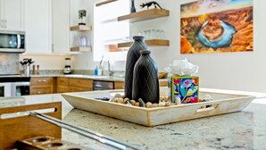 Een keuken met witte kasten, een magnetron en een houten plank. Op het aanrecht staat een dienblad met een vaas en een kleurrijke doos.