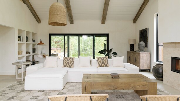 A modern living room with a white sectional sofa, a wooden coffee table, wicker chairs, a bookshelf, and a fireplace. - The Still House ~ Acreage · Hot Tub · Sauna · Gym (Bend)