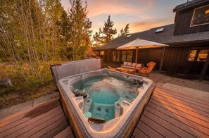 A hot tub on a wooden deck with a patio chair and umbrella, surrounded by trees and a building.