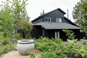 Ett modernt svart hus med ett sluttande tak, omgivet av en trädgård med olika växter och en stor planteringslåda i sten.
