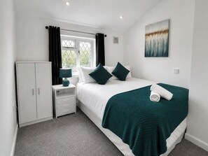 A bedroom with a bed, a wardrobe, a bedside table with a lamp, a window with curtains, and a painting on the wall. - 4 Bed Home Near Legoland | Sleeps 8 | Parking (Berkshire)