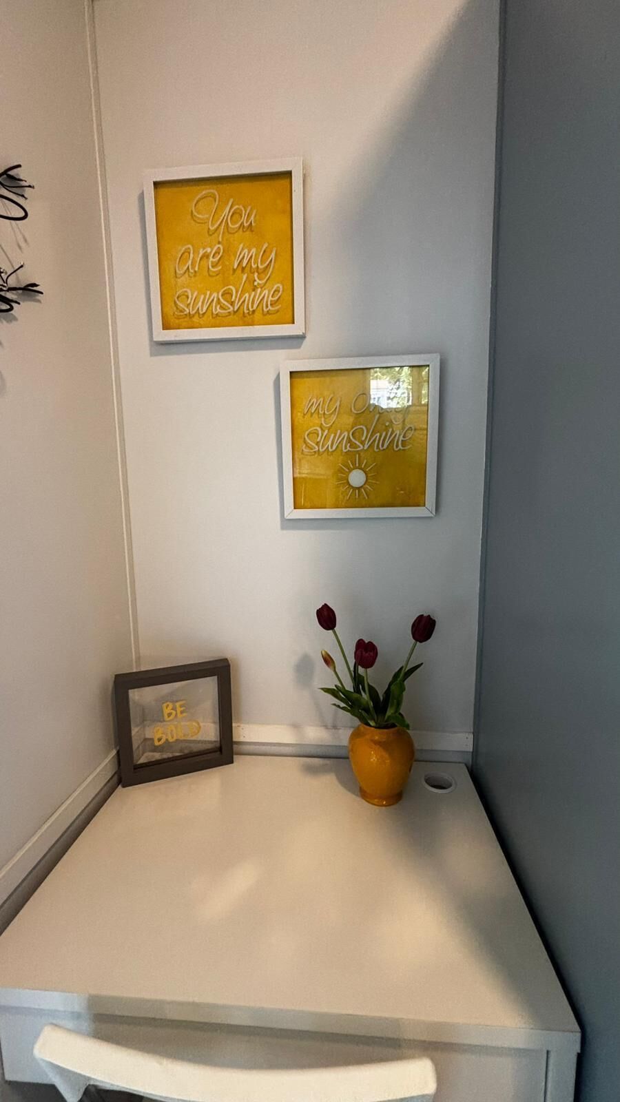 A corner of a room with a white desk, two framed inspirational quotes, a small vase with red flowers, and a framed picture with the text 'BE BOLD'.