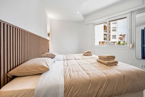Une chambre avec deux lits, une tête de lit en bois, une fenêtre donnant sur des immeubles et une porte bleue.